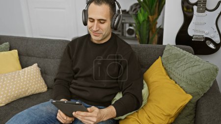 Foto de Hombre hispano sentado tranquilamente en un sofá en casa, usando auriculares y una tableta con una guitarra en el fondo. - Imagen libre de derechos