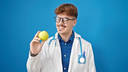 Téléchargez les photos : Jeune homme hispanique médecin souriant confiant tenant pomme verte sur fond bleu isolé - en image libre de droit