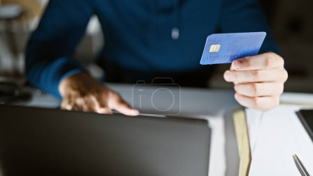 Hispanischer Mann mit Kreditkarte und Laptop im modernen Büroumfeld, der Online-Zahlungen oder Business-Banking darstellt.