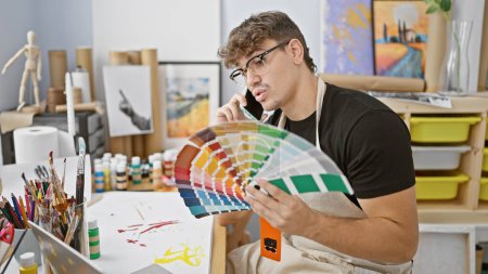 Photo for Charming young hispanic man, art student at college, fully absorbed in a vibrant palette's conversation via smartphone, choosing the perfect paint color in studio, a brush in hand. - Royalty Free Image