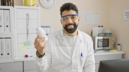 Un jeune hispanique souriant en blouse de laboratoire tenant une bouteille de médicament se tient dans une clinique avec du matériel en arrière-plan.