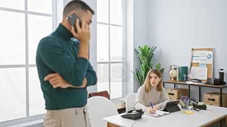 Foto de En una oficina, un hombre en una llamada telefónica se para mientras una mujer enfocada escribe en su escritorio, encarnando la colaboración en el lugar de trabajo. - Imagen libre de derechos