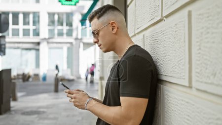 Foto de Hombre hispano joven guapo usando teléfono inteligente en la calle de la ciudad con los edificios borrosos fondo. - Imagen libre de derechos