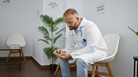 Foto de Joven guapo, un médico de cara seria, enviando mensajes de texto en el teléfono inteligente mientras se sienta relajadamente en la silla de la sala de espera de la clínica inmerso en su trabajo médico. - Imagen libre de derechos