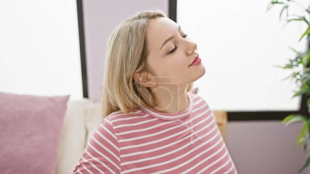 Téléchargez les photos : Jeune femme détendue aux cheveux blonds profitant d'un moment paisible à l'intérieur, assise à la maison. - en image libre de droit