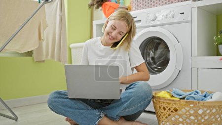 Photo for Young blonde woman using laptop talking on smartphone at laundry room - Royalty Free Image