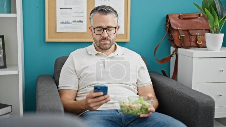 Photo for Grey-haired man business worker using smartphone eating salad at the office - Royalty Free Image