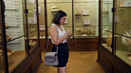 Téléchargez les photos : Jeune belle femme hispanique visitant exposition d'histoire naturelle à l'aide d'un smartphone au Musée d'histoire naturelle de Vienne - en image libre de droit