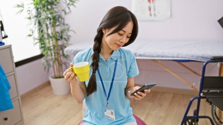 A smiling young asian woman nurse in a clinic, holding a cup and smartphone in a rehabilitation room.