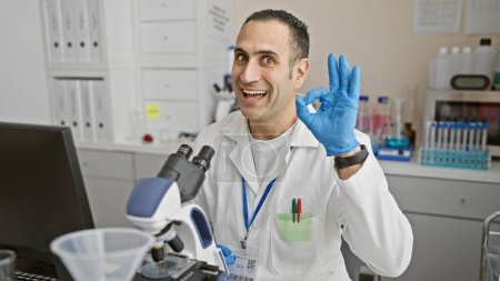 Foto de Hombre científico hispano con bata de laboratorio haciendo gestos de "bien" con microscopio y tubos de ensayo en laboratorio. - Imagen libre de derechos