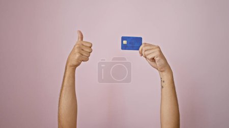 Photo for Hispanic man's arms giving thumbs up holding blue credit card isolated on pink background - Royalty Free Image