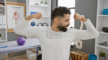 Photo for Handsome hispanic man flexing muscles with smile in rehab clinic interior. - Royalty Free Image