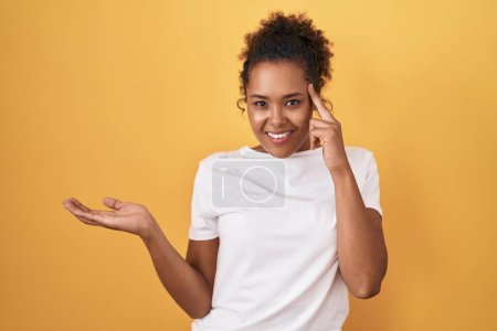 Photo for Young hispanic woman with curly hair standing over yellow background confused and annoyed with open palm showing copy space and pointing finger to forehead. think about it. - Royalty Free Image