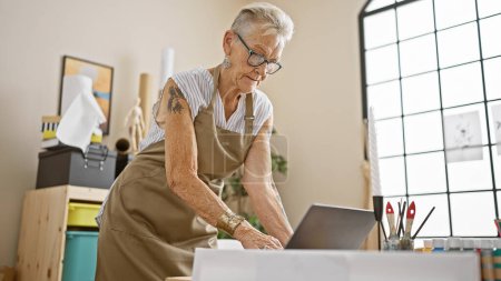 Photo for Confident grey-haired, short-haired senior woman artist joyfully embraces technology, using laptop for creativity in indoor art studio class - Royalty Free Image