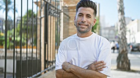 Photo for Smiling young hispanic man radiates confidence while casually standing with crossed arms - a joyful expression on sunny city street - Royalty Free Image