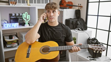 Photo for Handsome young hispanic male artist engrossed in a serious conversation on his smartphone at a classic music studio, his treasured classical guitar in hand - Royalty Free Image
