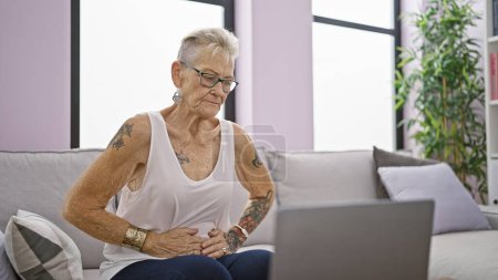 Photo for Serious grey-haired senior woman, focused on using laptop, sitting on sofa at home, touching stomach with an ache, sign of illness or sickness - Royalty Free Image