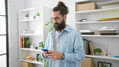 Photo for A bearded hispanic man in casual attire focused on his smartphone in a bright modern office. - Royalty Free Image