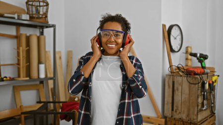 Téléchargez les photos : Femme souriante avec lunettes de sécurité et écouteurs dans une boutique de bois - en image libre de droit