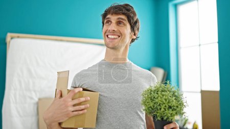Téléchargez les photos : Jeune homme hispanique souriant confiant tenant paquet et plante à la nouvelle maison - en image libre de droit