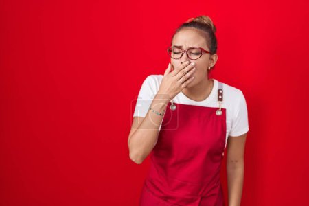 Foto de Mujer hispana joven con delantal de camarera sobre fondo rojo aburrida bostezando cansada cubriendo la boca con la mano. inquietud y somnolencia. - Imagen libre de derechos