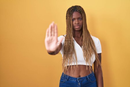 Foto de Mujer afroamericana con el pelo trenzado de pie sobre fondo amarillo haciendo dejar de cantar con la palma de la mano. expresión de advertencia con gesto negativo y grave en la cara. - Imagen libre de derechos