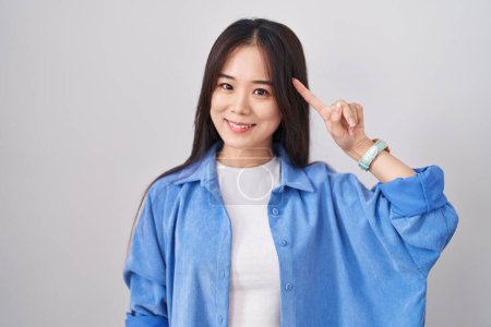 Téléchargez les photos : Jeune femme chinoise debout sur fond blanc souriant pointant vers la tête avec un doigt, bonne idée ou pensée, bonne mémoire - en image libre de droit
