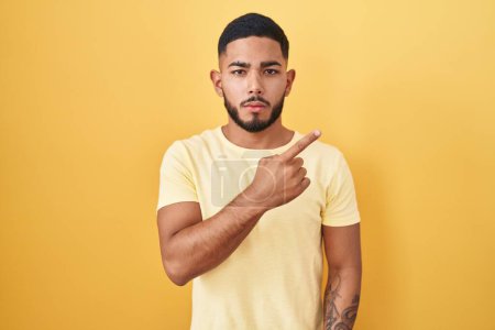 Téléchargez les photos : Jeune homme hispanique debout sur fond jaune pointant avec le doigt de la main sur le côté montrant la publicité, visage sérieux et calme - en image libre de droit