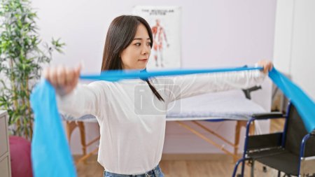 Photo for A young asian woman exercises with a resistance band in a bright rehabilitation clinic room. - Royalty Free Image