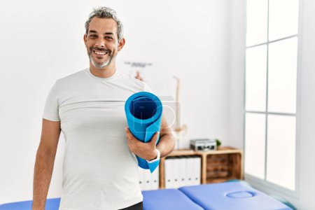 Téléchargez les photos : Homme moyen aux cheveux gris patient en désintox tenant un tapis de yoga à la clinique de désintox - en image libre de droit