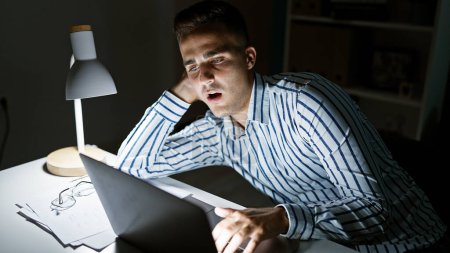 Schöner hispanischer Mann gähnt in gut beleuchtetem Büro während der späten Arbeitszeit und zeigt Müdigkeit