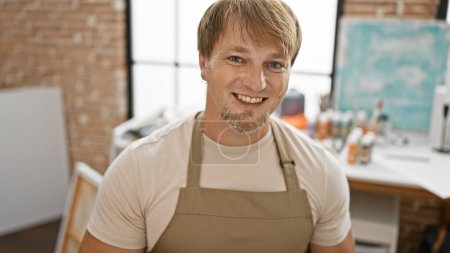 Photo for Portrait of a smiling caucasian man with a blond beard wearing an apron in an art studio setting. - Royalty Free Image
