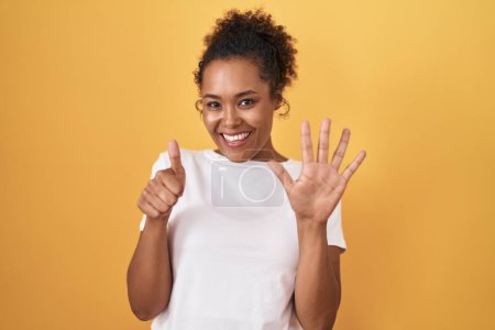 Téléchargez les photos : Young hispanic woman with curly hair standing over yellow background showing and pointing up with fingers number six while smiling confident and happy. - en image libre de droit