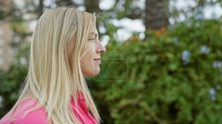 Téléchargez les photos : Profil d'une femme blonde réfléchie dans une veste rose en plein air par la verdure dans un cadre de parc. - en image libre de droit