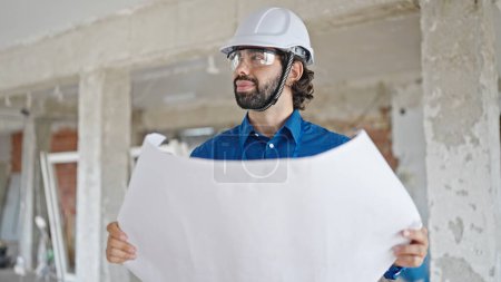 Photo for Young hispanic man architect holding blueprints looking around at construction site - Royalty Free Image