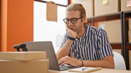 Photo for Focused man with glasses working on a laptop in a warehouse surrounded by cardboard boxes. - Royalty Free Image