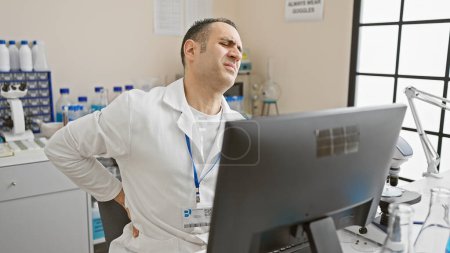 Foto de Un hombre con una bata de laboratorio expresando molestias mientras trabajaba en un laboratorio médico en interiores. - Imagen libre de derechos