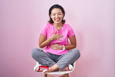 Foto de Mujer joven hispana sentada en silla sobre fondo rosa sonriendo y riendo en voz alta porque divertida broma loca con las manos en el cuerpo. - Imagen libre de derechos