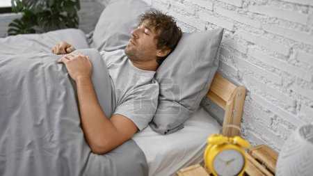 Foto de Hombre hispano durmiendo pacíficamente en un moderno dormitorio de ladrillo blanco, con un reloj despertador amarillo en primer plano. - Imagen libre de derechos