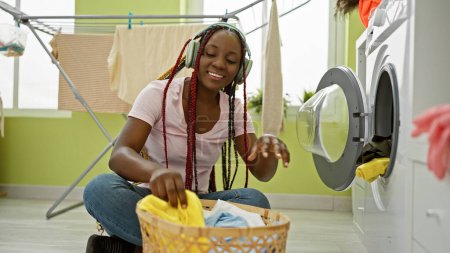 Photo for Joyous african american woman listening to soulful music, washing clothes with a gleaming smile in a cozy laundry room - Royalty Free Image