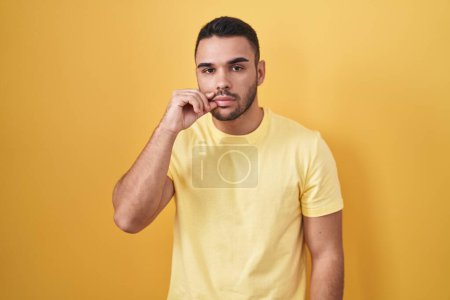 Téléchargez les photos : Jeune homme hispanique debout sur fond jaune bouche et lèvres fermées comme zip avec les doigts. secret et silencieux, tabou parlant - en image libre de droit