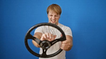 Handsome young man feigning driving with intense expression against a plain blue background.