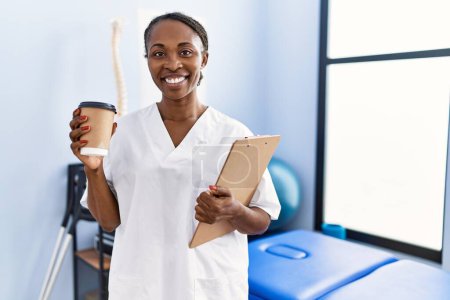 Foto de African american woman physiotherapist holding medical report drinking coffee at rehab clinic - Imagen libre de derechos