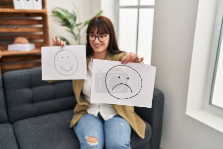 Photo for Young beautiful hispanic woman psychologist holding sadness and smiling emoji papers at psychology clinic - Royalty Free Image