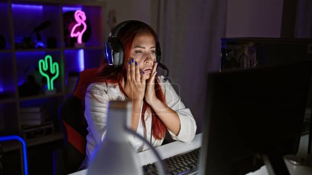 Photo for Overworked and stressed young redhead female streamer, battling stress while streaming a game from her gaming room late at night - Royalty Free Image