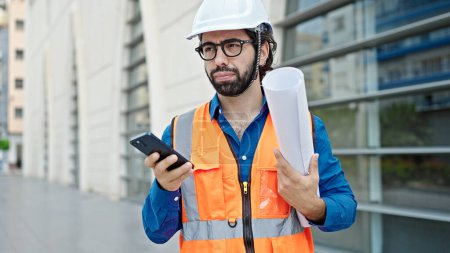 Photo for Young hispanic man architect holding blueprints using smartphone at construction place - Royalty Free Image