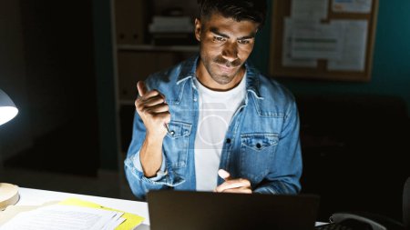 Hispanic man working late in an office, illuminated by a desk lamp and looking at his laptop with a focused expression.