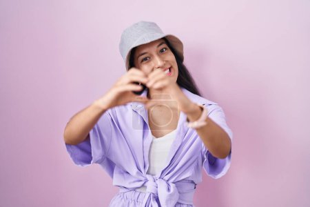 Foto de Mujer hispana joven de pie sobre fondo rosa con sombrero sonriendo en el amor haciendo forma de símbolo del corazón con las manos. concepto romántico. - Imagen libre de derechos