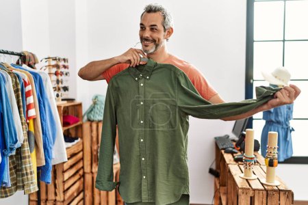Foto de Hombre de pelo gris de mediana edad cliente sonriendo confiado sosteniendo la camisa de rack en la tienda de ropa - Imagen libre de derechos