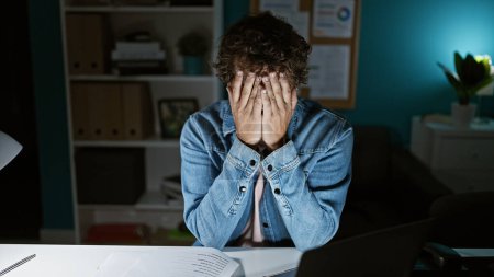 Photo for A stressed man covers his face with hands at his desk late at night in the office, reflecting overwork and fatigue. - Royalty Free Image